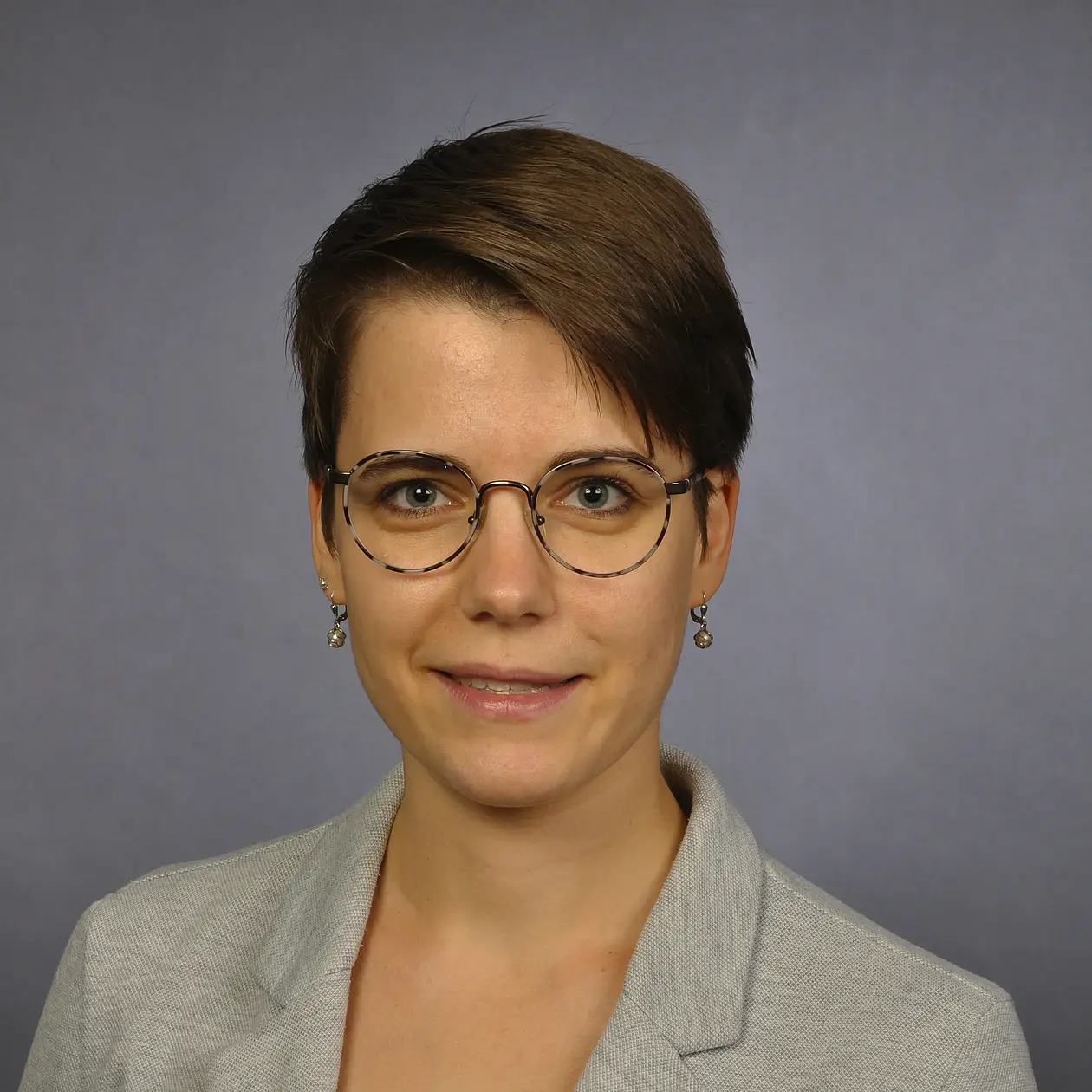 A portrain photo of Annika Thiem, a white woman with short brown hair swept to the left side, wearing glasses and a light grey blazer jacket facing the camera.